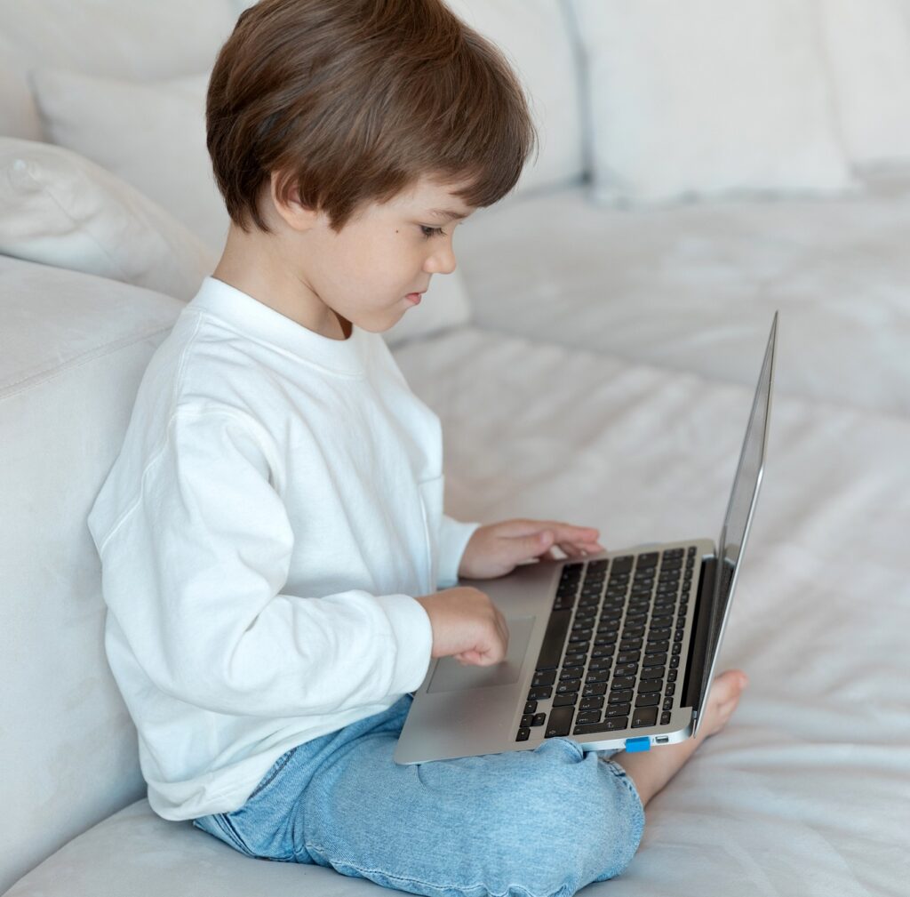 A preschool kid uses lap top, sitting on the sofa